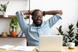 © fizkes - Smiling Afro-American businessman holding hands behind head sitting at office