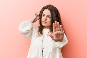 Wall Mural - Young plus size curvy woman listening music with headphones standing with outstretched hand showing stop sign, preventing you.