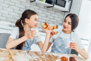 Beautiful woman and her cute little daughter in aprons are looking at camera  and smiling while tasting their muffins and drinking milk!