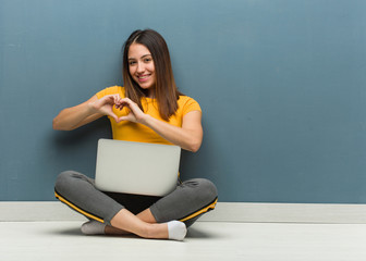 Young woman sitting on the floor with a laptop doing a heart shape with hands