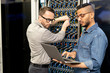 © Seventyfour - Serious young multi-ethnic IT engineers in casual shirts standing in database center and setting up computer cluster while examining information on laptop
