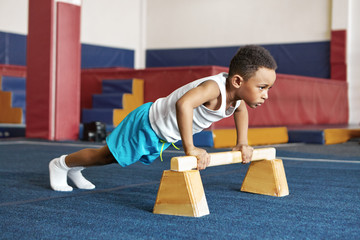 Wall Mural - Sports, motivation and strength concept. Indoor image of serious disciplined dark skinned black child doing push ups during cross fit workout using two wooden parallel bars, having focused look