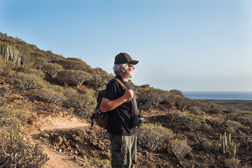 Senior people hiking in the mountain and stops to look the landscape with a binoculars. Happy excursion outdoor. One caucasian adult. Volcanic landscape and blue sky