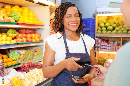 Cuadros en Lienzo Smiling pretty young grocery store worker with card reader talking to customer