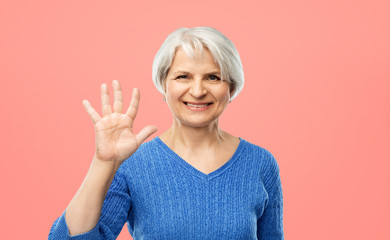 Wall Mural - gesture and old people concept - portrait of smiling senior woman in blue sweater showing palm or five fingers over pink or living coral background