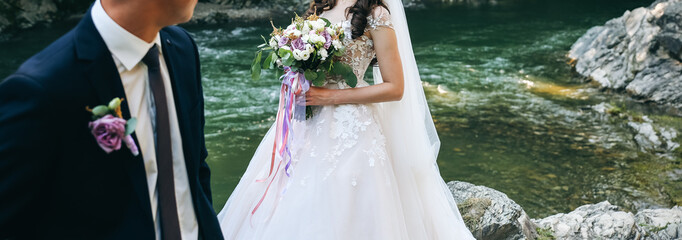 Wall Mural - Wedding couple is hugging near the mountain river. Groom and bride with a bouquet in hands. Beautiful flowers. Nature waterfall.