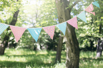 Pastel bunting flags hanging among trees. Summer garden party. Outdoor birthday, wedding decoration. Midsummer, festa junina concept. Selective focus. Natural blurred background, sunny haze.