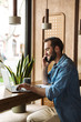 © Drobot Dean - Photo of young smiling man talking on cellphone with laptop and clipboard while working in cafe indoors