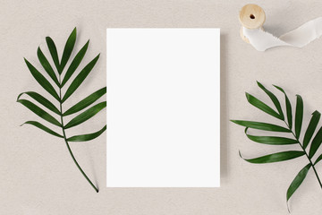 Feminine wedding stationery mock-up scene. Blank greeting card, green palm leaves and silk ribbon on beige textured table background. Tropic summer styled photo, web banner. Flat lay, top view.