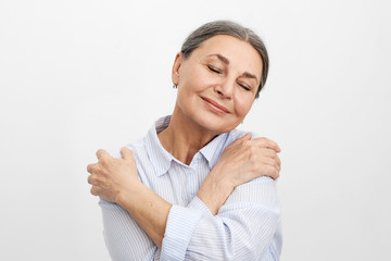 Wall Mural - Self-love, joy and happiness concept. Portrait of happy joyful elderly mature lady with wrinkled skin and gray hair posing in studio, dressed in blue shirt, closing eyes and embracing herself