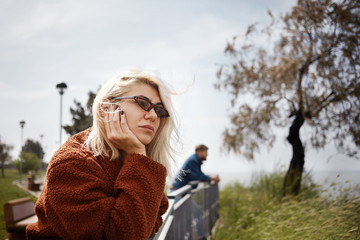 Picture of trendy looking young woman in shades having frustrated disappointed facial expression after quarrel with her boyfriend who’s standing in background. People and relationships concept