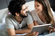 © NDABCREATIVITY - Happy young couple looking a tablet together and laughing while lying on the bed