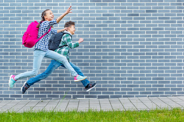 two happy schoolchildren with backpacks run to school on street next to an brick wall. cheerful cute