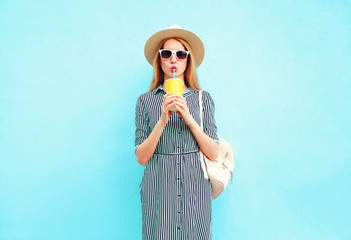 Wall Mural - Young woman drinking fruit juice from cup, wearing summer round straw hat, striped dress on blue wall background