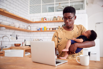 Wall Mural - Multi-Tasking Father Holds Sleeping Baby Son And Works On Laptop Computer In Kitchen