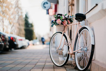 White Bicycle With Basket Of Flowers Standing Near The Door On The Street In City.