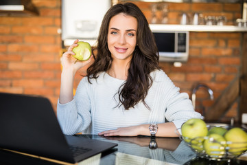 Portrait of a beautiful young student girl with apple sitting indoors using laptop computer.