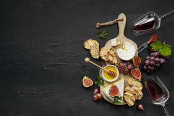 Wall Mural - Two glasses of red wine and a tasty cheese plate with fruit, grape and toasted bread on a wooden kitchen plate on the black stone background, top view, copy space. Gourmet food and drink.