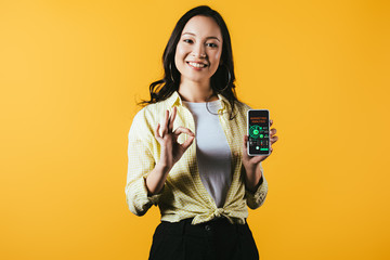 Wall Mural - smiling asian girl showing ok sign and smartphone with marketing analysis, isolated on yellow