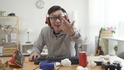 Wall Mural - nervous asian man using desktop computer wear headset at home playing online video game. intense male talking on headphones with enemy on internet. teenage indoorsy at messy home on summer holidays.