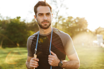 Wall Mural - Handsome young strong sports man posing outdoors at the nature park location with skipping rope.