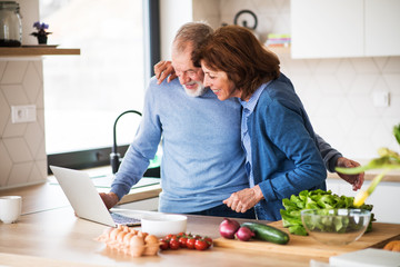 Wall Mural - A portrait of senior couple with laptop indoors at home, cooking.