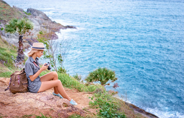 Wall Mural - Photo shooting and traveling. Young woman in hat with rucksack holding camera enjoying sea view.