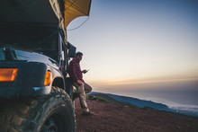 Traveler People With Car And Camping Concept - Lonely Man Use Cellular Phone To Connect To Internet Outside His Vehicle - Mountain And Nature Outdoor Around - Enjoying Freedom And Alternative Vacation