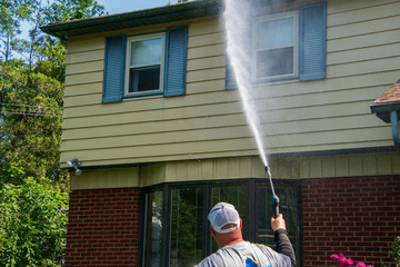 One heavy overweight Caucasian man spraying water solution on the front of a house as part of his pressure washing service.