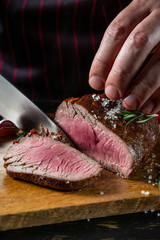 Slicing juicy beef steak by knife in chef hands closeup. Food cooking concept. Dark black background copy space.