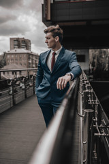 Wall Mural - Portrait of handsome fashion businessman model dressed in elegant suit posing on street background. City urban style. Success lifestyle