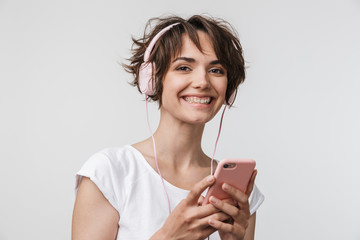 Poster - Young pretty excited happy woman posing isolated over white wall background using mobile phone listening music with earphones.