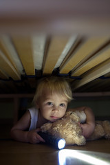 Poster - Little child, hiding under the bed, hugging teddy bear and holding flashlight,