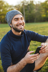 Wall Mural - Portrait of handsome sporty man using smartphone and sitting on grass during workout in green park