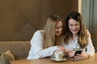 © Alvaro Hernandez/ADDICTIVE STOCK - Two stylish female friends with cups of fresh coffee smiling and looking at phone while sitting at table in cozy restaurant