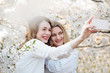 © Alvaro Hernandez/ADDICTIVE STOCK - Two cheerful young women smiling and taking selfie near trees with white flowers on sunny day in park