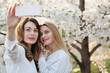 © Alvaro Hernandez/ADDICTIVE STOCK - Two cheerful young women smiling and taking selfie near trees with white flowers on sunny day in park