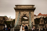 Fototapeta Boho - Young beautiful stylish pair of newlyweds on a bridge in Budapest, Hungary. Beautiful woman in a white wedding dress and handsome man in suit.