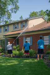 Man pressure washes the siding on his house with his two teenage sons watching. The man is spraying water on the top floor. The house has blue shutters