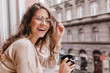 © Look! - Close-up portrait of laughing brunette girl in beige sweater drinking coffee on city background. Lovable lady standing near window and looking at beautiful building with cup of tea.
