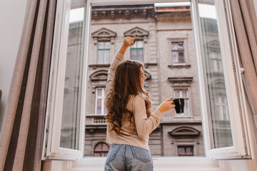 Wall Mural - Indoor photo from back of trendy woman in jeans drinking tea and looking at city. Inspired female model with curly long hair stretching near window.