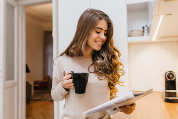 Wall Mural - Stylish white woman with happy smile reading magazine and holding cup of latte. Indoor photo of glamorous long-haired girl drinking coffee in good weekend morning.