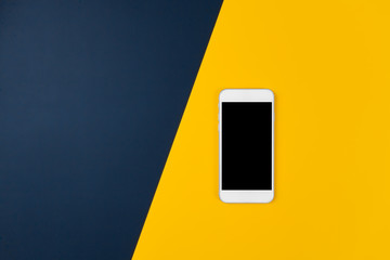 A white mobile phone on yellow and denim blue background. Top view with copy space. Selective focus.