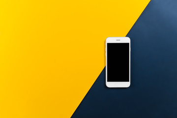 A white mobile phone on yellow and denim blue background. Top view with copy space. Selective focus.