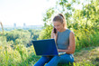 © broniktav - The girl is talking on a smartphone and looking at the laptop monitor. Girl in jeans in nature with a laptop. Doing business while on holiday.