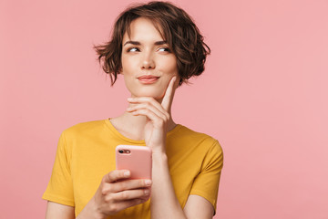 Poster - Thinking dreaming young beautiful woman posing isolated over pink wall background using mobile phone.