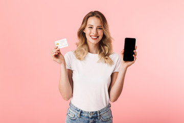Young blonde woman posing isolated over pink wall background holding credit card using mobile phone showing display.
