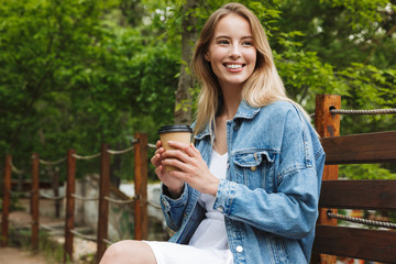 Poster - Amazing happy young woman student posing outdoors in park drinking coffee.