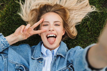 Poster - Excited happy young woman posing outdoors in park lies on grass take a selfie.