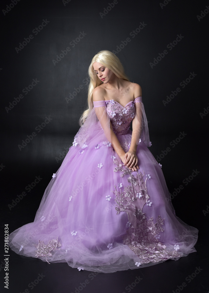full length portrait of a blonde girl wearing a fantasy fairy inspired  costume, long purple ball gown with fairy wings, sitting pose on a dark  studio background. Stock Photo | Adobe Stock
